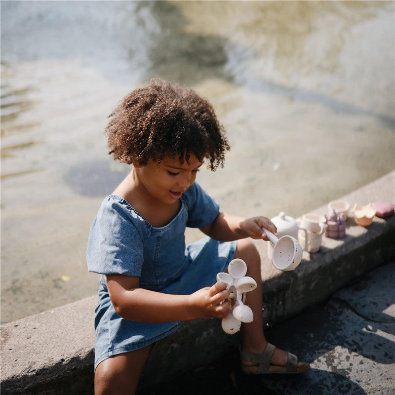 A child plays with the Mushie Water Wheel Bath Toy by experimenting with small white water scoops near a pond.