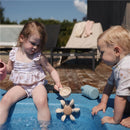 A young girl in a floral swimsuit plays with a Mushie Water Wheel Bath Toy in a pool, while a toddler watches.