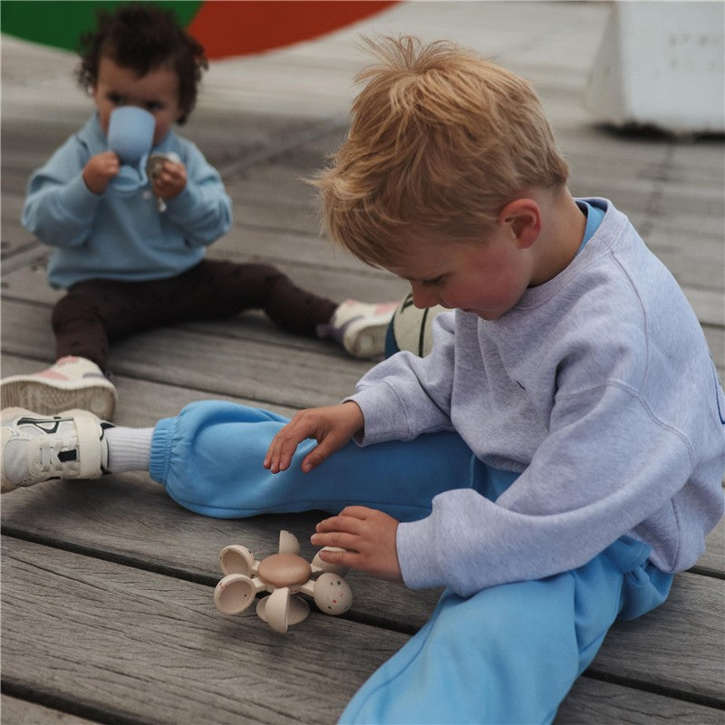 Child playing with the Mushie Water Wheel Bath Toy on wooden flooring, while another child drinks from a cup in the background.