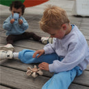 Child playing with the Mushie Water Wheel Bath Toy on wooden flooring, while another child drinks from a cup in the background.
