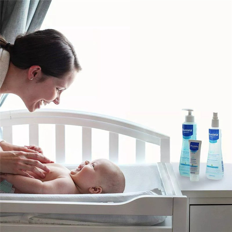 Mother smiling at her baby in a crib, with Mustela Hair Styler & Skin Freshener products displayed on a nearby surface.