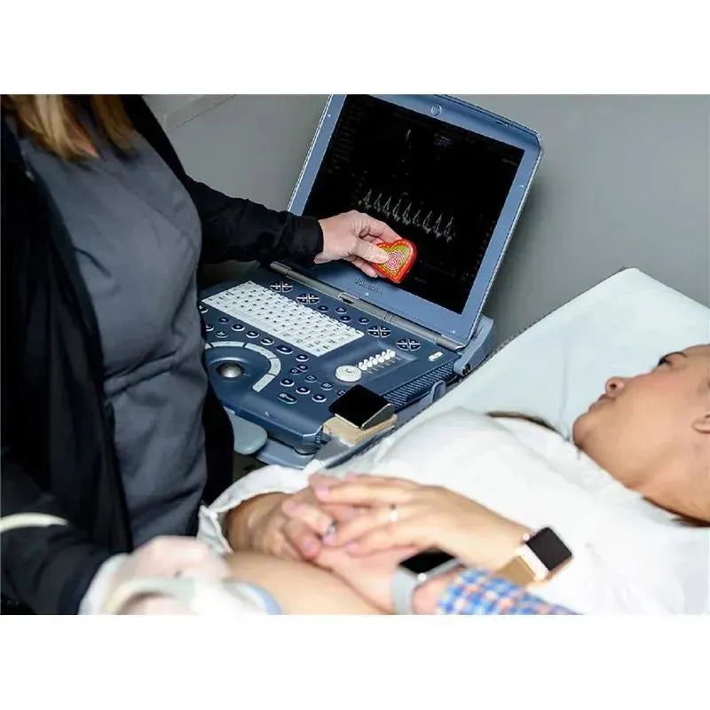 A woman lies on a medical bed while a technician uses a device displaying a heartbeat pattern, holding a heart-shaped object.