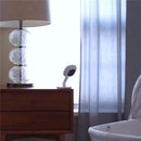 Nanit Flex Multi-Stand baby monitor accessory on a nightstand beside a decorative lamp and a baby bassinet.