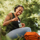Smiling woman in a tank top using Natrapel tick and insect repellent wipes while sitting outdoors beside an orange backpack.