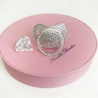Crystal-embellished Neonilla orthodontic pacifier on a pink circular box, featuring a diamond-shaped logo.