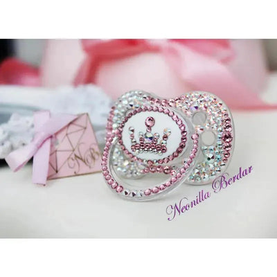 Pink bling pacifier featuring a crown design, adorned with sparkling crystals and rhinestones, with a gift box nearby.