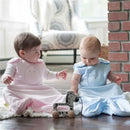 Two infants wearing a pink and a blue Nested Bean Zen Sack sit on a rug, playing with a toy zebra in a bright room.