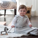 A baby wearing a solid gray Nested Bean Zen Sack sits on a white blanket, holding a wooden zebra pull toy.