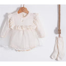 Knitted cream baby outfit with ruffled collar and sheer overlay, accompanied by matching socks hanging nearby.