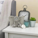 Gray plush elephant with large ears holding a pacifier, set on a table with a clock, succulent plant, and jar of blue beads.