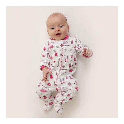 Baby girl in a zipped footie covered in cosmetics patterns, smiling and posing against a light background.
