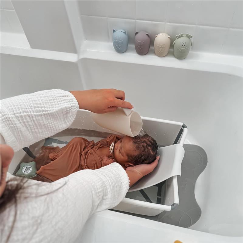 A caregiver gently pours water from a Nouka Bath Rinse Jug onto a baby in a bath support in a bathtub.