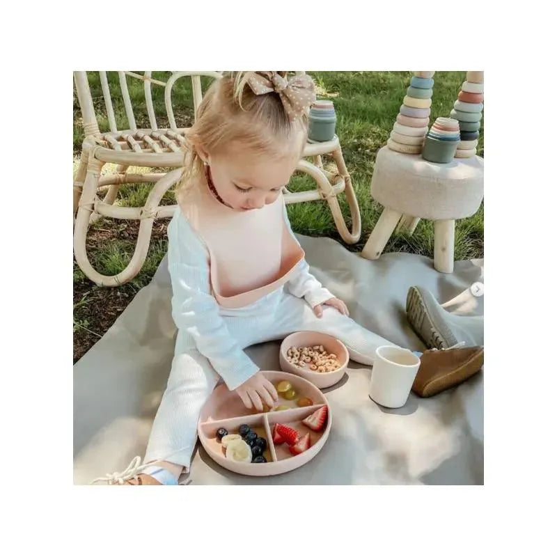 Toddler sitting on a blanket, using a pink divided suction plate filled with fruits, alongside a bowl of cereal and a cup.