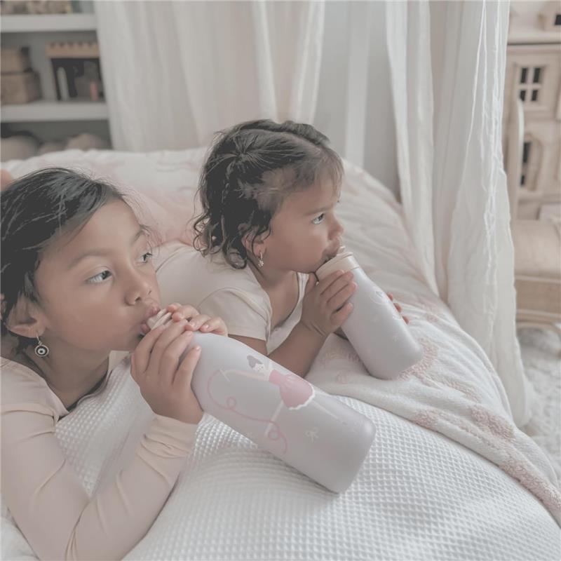Two young girls seated on a bed, each holding a light purple Nouka insulated stainless steel water bottle featuring a ballerina design.