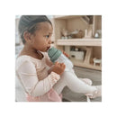 Child in a soft pink outfit sipping from a Nouka non-spill straw cup with a green lid, seated on a light-colored surface.