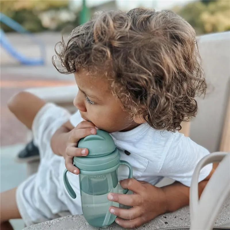 Child with curly hair holding a Nouka Straight Straw Cup in Soft Sand, looking intently at the cup while sitting outdoors.