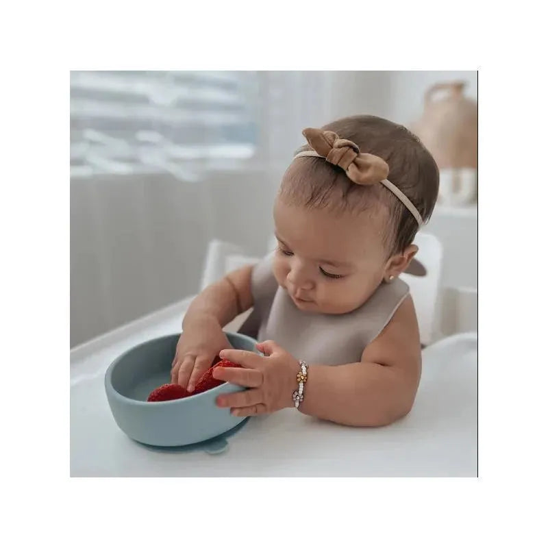 Baby in a high chair reaching into a blue suction bowl, playing with red strawberries, wearing a beige bib and headband.