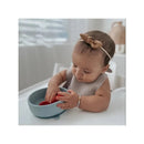 Baby in a high chair reaching into a blue suction bowl, playing with red strawberries, wearing a beige bib and headband.