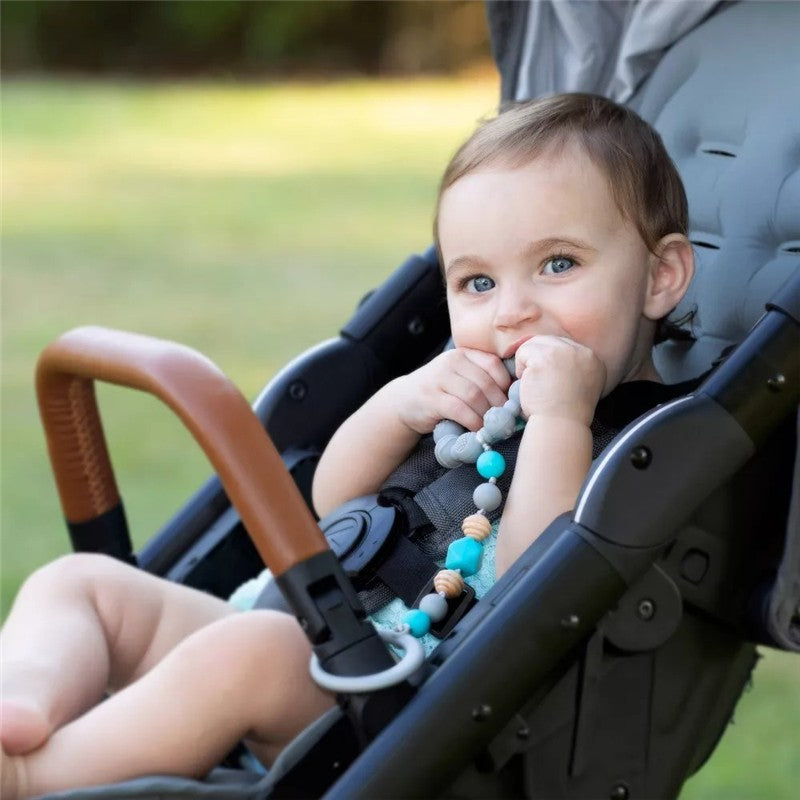 A baby in a stroller chews on a gray Nuby Tag A Long Teether, featuring colorful beads and a secure attachment.