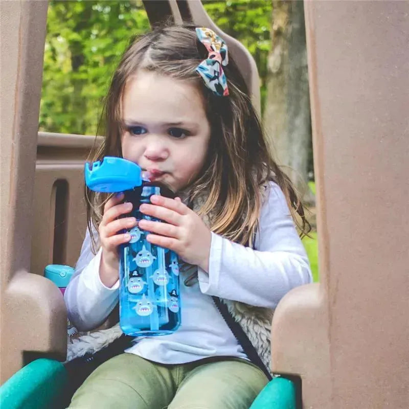 A child with long hair and a patterned bow drinks from a blue Nuby water bottle decorated with shark prints.