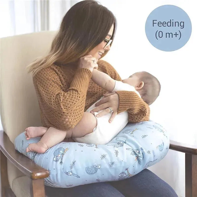 Nuby Dr Talbots white nursing pillow supports a mother feeding her infant, featuring a blue cover with whimsical animal designs.