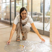 A woman kneels on a floor, placing a transparent Nuby waterproof floor mat down, with small food pieces scattered on it.