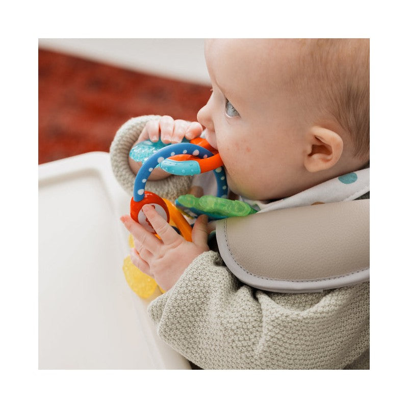 A baby holds colorful Nuby Ice Gel Teether Keys, chewing on the blue and orange ring while seated in a high chair.