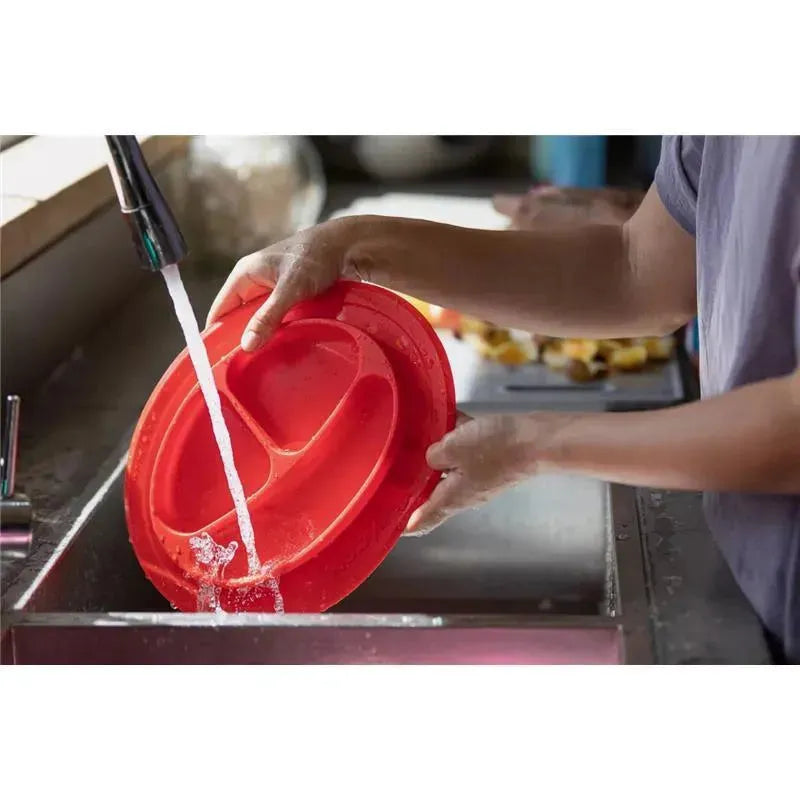 Sectioned red silicone feeding mat being rinsed under running water in a kitchen sink.