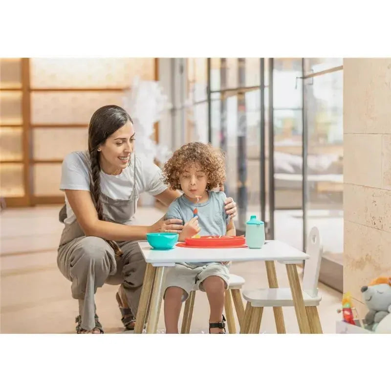 A mother assists a child at a table with a Nuby sectioned silicone feeding mat, featuring vibrant food in bowls.