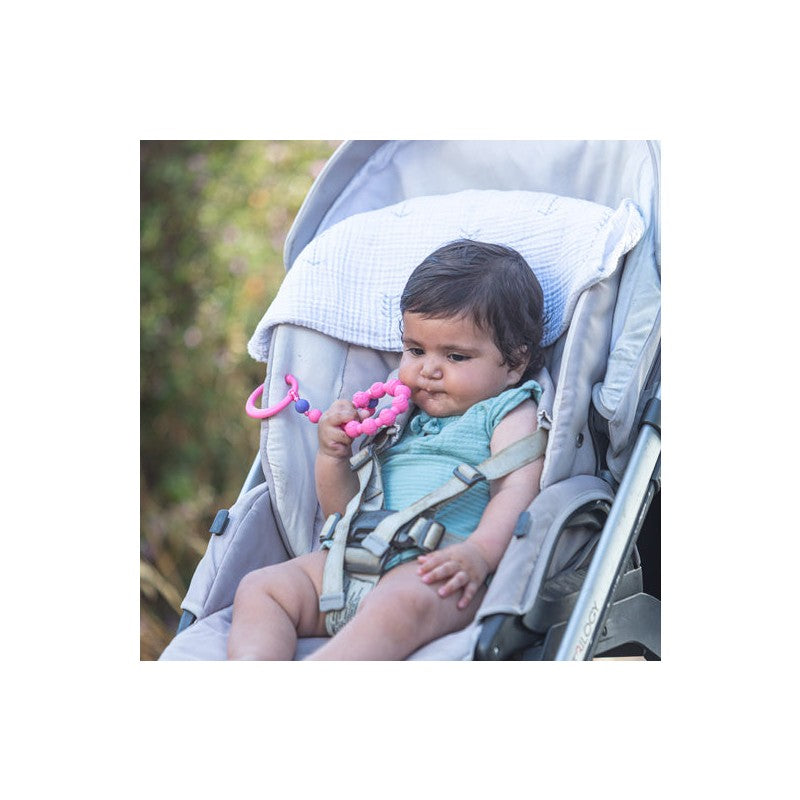 A baby in a stroller holds a Nuby Tag A Long Teether in pink, looking curious while sitting comfortably.