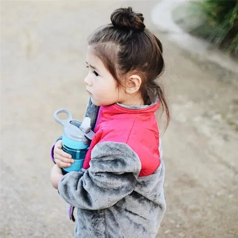 Young girl holding a Nuby Thirsty Kids Flip It Reflex bottle in aqua while wearing a gray and pink jacket.
