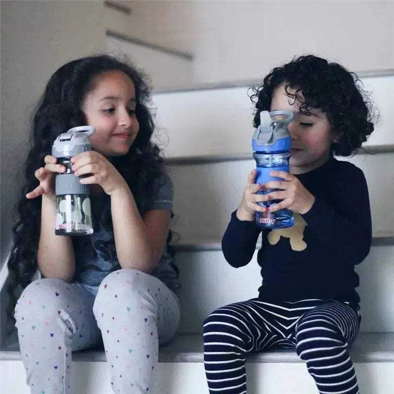 Two children sit on stairs, each holding a Nuby Thirsty Kids Flip It Reflex water bottle, one grey and clear, the other blue.
