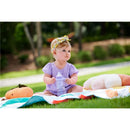 A toddler wearing a purple outfit and headband sits on a colorful blanket outdoors, holding a clear sippy cup.