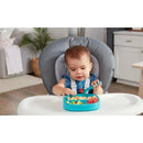 A baby seated in a high chair uses a blue Nuk Pretensil Dipper Spoon to eat from a divided tray with colorful food.