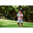 Toddler in a pink shirt and shorts joyfully running on grass, holding a blue Nuk sippy cup.