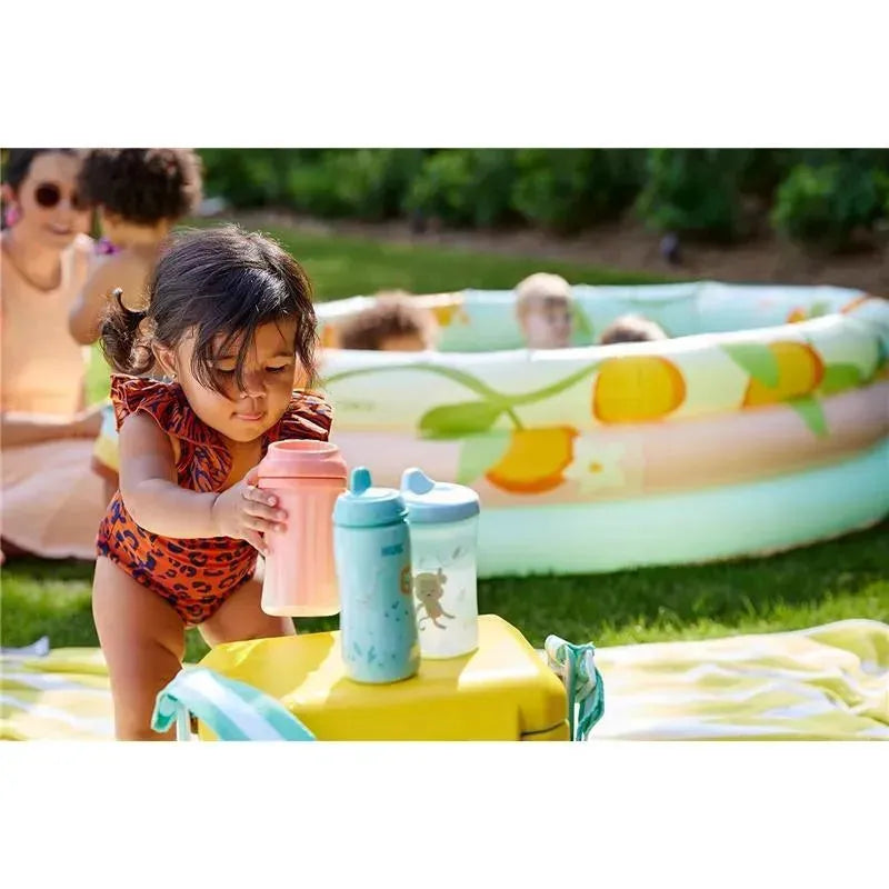 A toddler in a leopard-print swimsuit holds a pink sippy cup, with two additional sippy cups nearby, in a sunny outdoor setting.