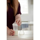 A person holds a Nuk Simply Natural baby bottle filled with milk, preparing to attach a nipple while a spare nipple sits nearby.