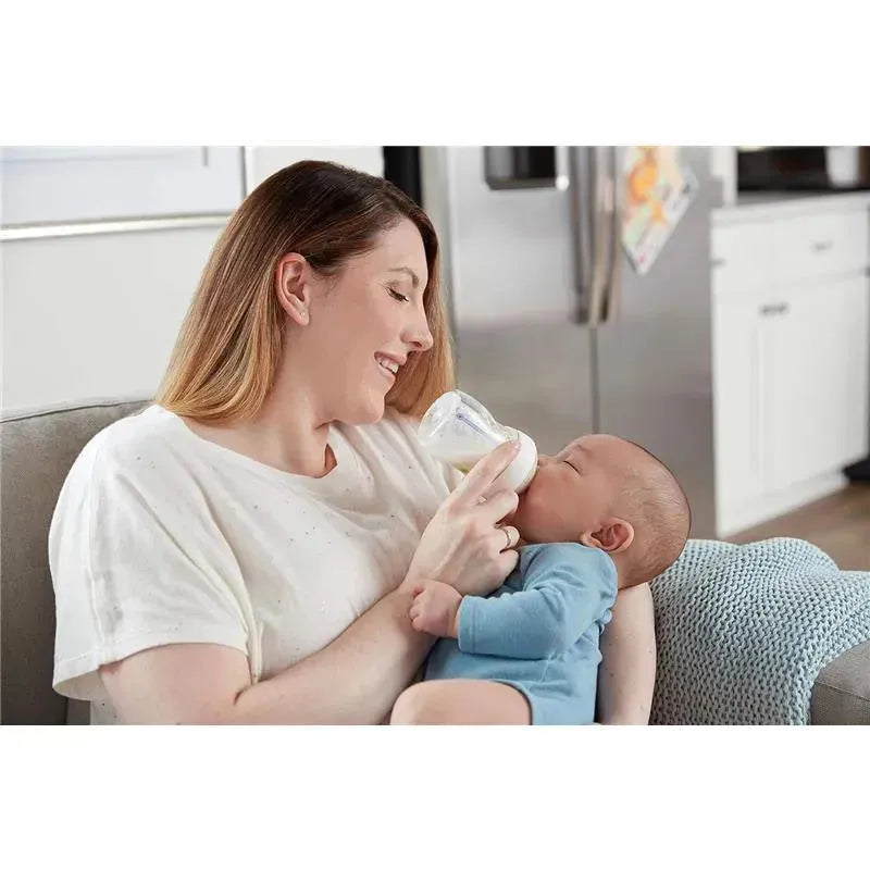 A woman smiles while feeding a baby with a Nuk Simply Natural baby bottle in a cozy living room setting.
