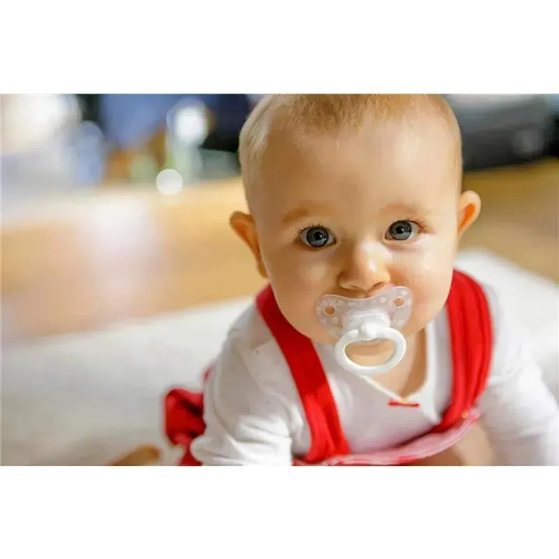 Baby in a red outfit with a white top and pacifier, sitting on a floor, smiling at the viewer.