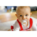 Baby in a red outfit with a white top and pacifier, sitting on a floor, smiling at the viewer.