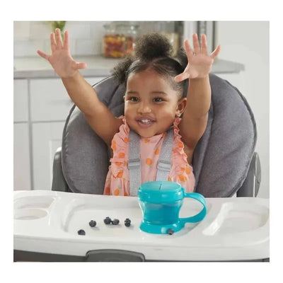 Toddler with raised hands sits in a high chair excitedly beside a turquoise NUK baby food storage container and blueberries.