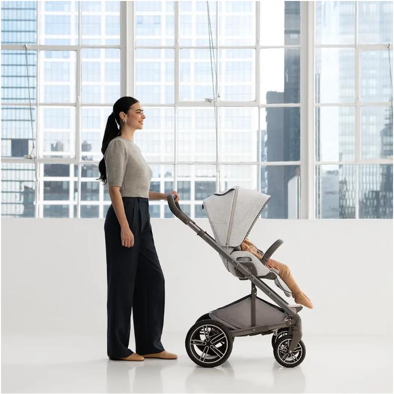 Nuna MIXX™ next Stroller in Sterling color, pushed by a smiling woman, with a child seated inside, set against a bright background.