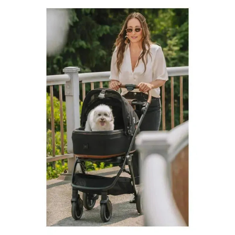 Woman pushing a Nuna Pet Maeve stroller with a small white dog inside, featuring a sleek black design and urban setting.