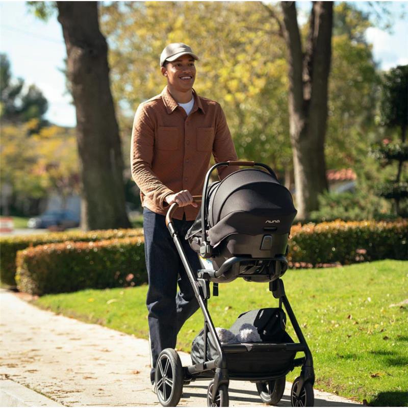 Adult man smiling while walking with a Nuna PIPA aire infant car seat in a stroller, outdoors in a park setting.