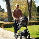 Adult man smiling while walking with a Nuna PIPA aire infant car seat in a stroller, outdoors in a park setting.