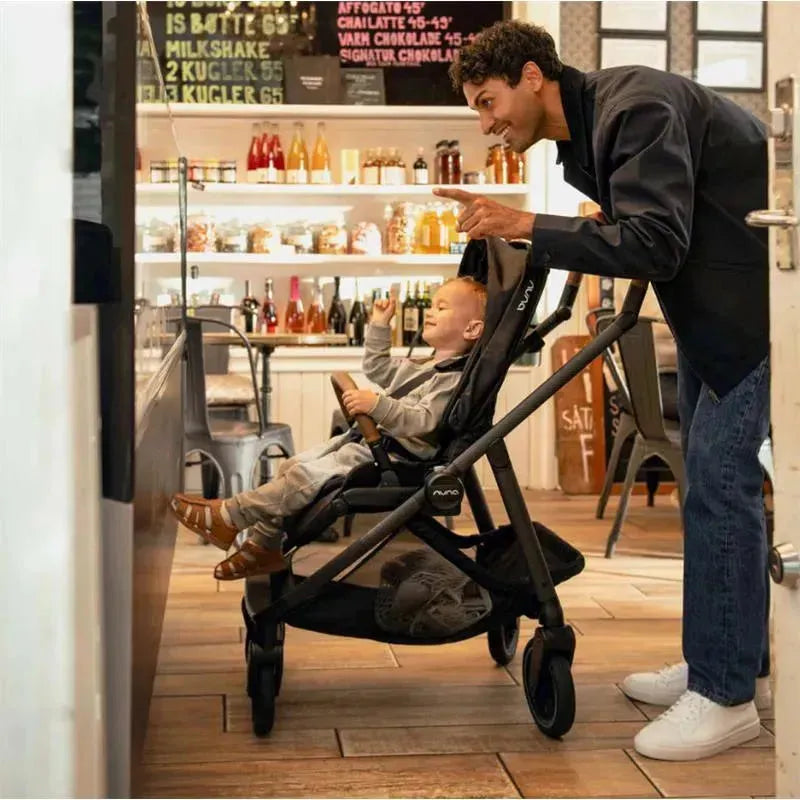 Nuna SWIV™ Stroller in Biscotti, featuring a child sitting excitedly while an adult gestures nearby in a café setting.