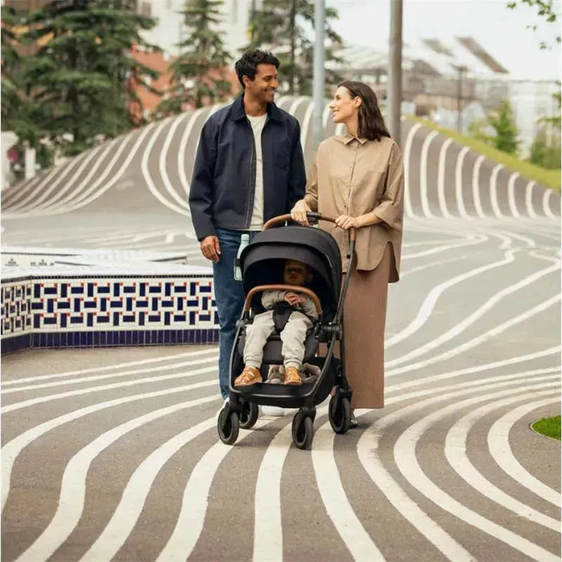 Nuna SWIV™ Stroller in Biscotti, with a child seated, as a couple strolls on a patterned walkway in an outdoor setting.