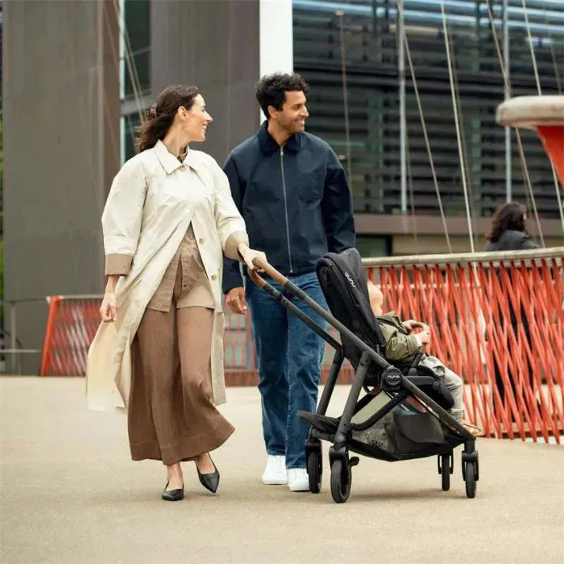 A couple walks on a path with a Nuna SWIV™ Stroller in Biscotti color, featuring a child seated and smiling.