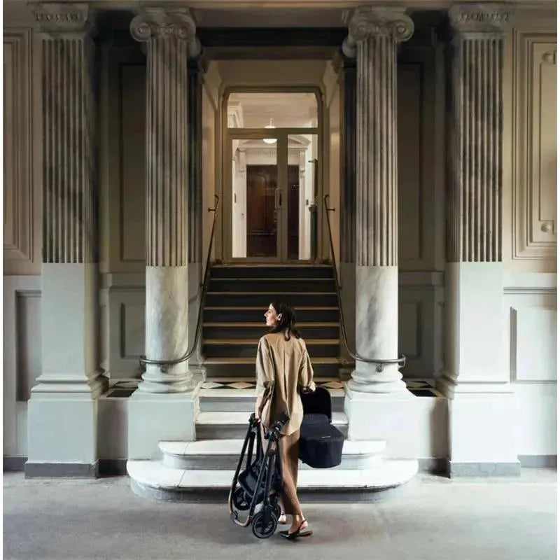 A woman carries a Nuna SWIV™ stroller in Chai color and a baby carrier while walking up steps in a grand indoor space.