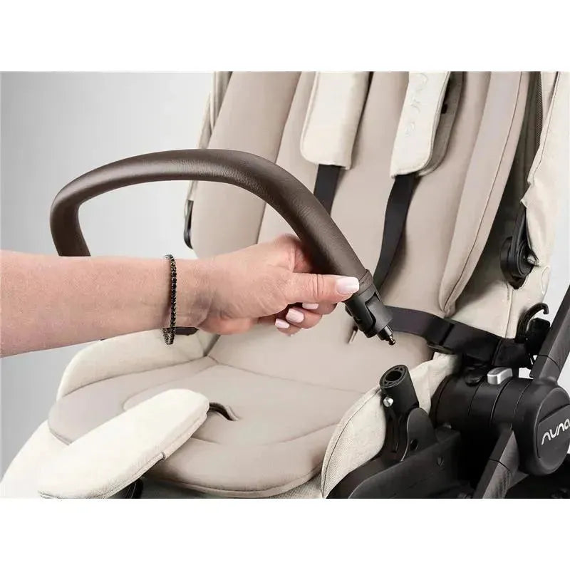 Nuna SWIV™ stroller in Chai color; a hand adjusts the brown leather handle on the seat with soft fabric.